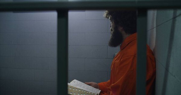 Male prisoner in orange uniform sits on the bed, reads Bible, looks at barred window in prison cell. Criminal serves imprisonment term for crime in jail. Detention center or correctional facility.