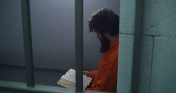 Male prisoner in orange uniform sits on the bed, reads Bible, looks at barred window in prison cell. Criminal serves imprisonment term for crime in jail. Detention center or correctional facility.