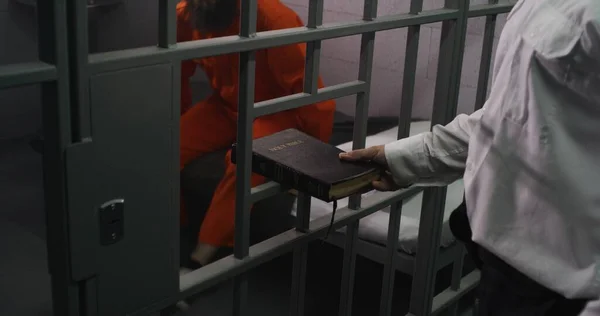 刑務所の警官はオレンジ色の制服を着た男性捕虜に聖書を贈った 刑事さんは刑務所のベッドに座って読んでいます 犯罪者は刑務所で有罪判決を受ける 拘留センター 神の概念に対する信仰 — ストック写真