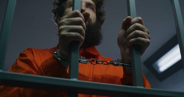 Prisoner in orange uniform and handcuffs holds metal bars, stands in prison cell. Criminal serves imprisonment term for crime in jail. Tired inmate in detention center. Justice system. Rack focus.