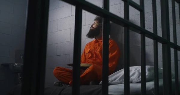 オレンジ色の制服を着た男性は ベッドに座り 聖書を読み 刑務所の監獄の窓を見ます インメイトは刑務所で有罪判決を受けた 矯正施設について 神の概念に対する信仰 — ストック写真