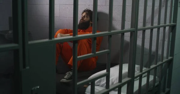 オレンジ色の制服を着た男性は ベッドに座り 聖書を読み 刑務所の監獄の窓を見ます インメイトは刑務所で有罪判決を受けた 矯正施設について 神の概念に対する信仰 — ストック写真
