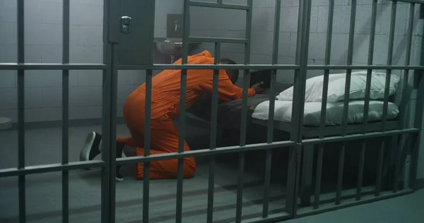 ベッドの近くでオレンジ色の制服を着た宗教的なアフリカ系アメリカ人の囚人は 聖書を保持する刑務所で神に祈ります 男性犯罪者は刑務所や拘置所で投獄期間を過ごしている 神への信仰 — ストック写真