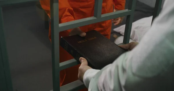刑務所の警官はオレンジ色の制服を着た男性捕虜に聖書を贈った 刑事さんは刑務所のベッドに座って読んでいます 犯罪者は刑務所で有罪判決を受ける 拘留センター 神の概念に対する信仰 — ストック写真