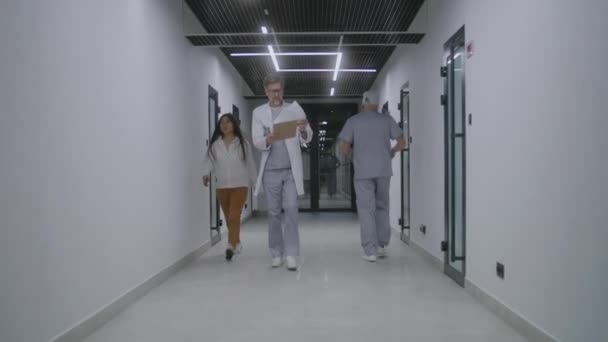 白い実験室のコートの成熟した医師は明るい診療所の廊下に沿って歩き 医学歴 診断または検査結果の論文を見ます 病院や医療センターの医療スタッフと患者 — ストック動画