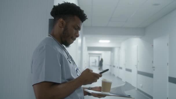 非裔美国人医疗专家站在医院走廊的侧壁上 医生和病人聊天或者用电话上网 在现代诊所或医疗中心工作的医务人员 — 图库视频影像