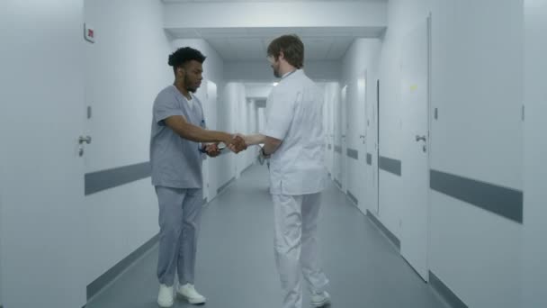 身穿白色制服的医生走过医疗中心走廊 与非裔美国人同事打招呼 并与女医生交谈 医院走廊里的多族裔医务人员 跟踪目标背景色 — 图库视频影像