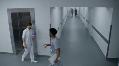 Yoğun klinik koridoru: Çeşitli doktorlar, sağlıkçılar, hemşireler, cerrahlar ve hastalar yürüyor. Çok ırklı doktorlar asansörü kullanıyor. Tıp personeli modern hastane ya da tıbbi tesiste çalışıyor. Yüksek açı.