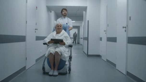 若い医師は 車椅子の上のシニア患者を医療キャビネットに運びます 古い女性はデジタルタブレットコンピュータを手に持っています 成熟した医師は同僚のための医療室のドアを開きます 職場の医療スタッフ — ストック動画