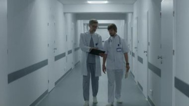 Sıhhiyeciler klinik koridorunda yürürler, iş tartışırlar. Olgun doktor MRI ve CT beyin taraması görüntülerini dijital tablet kullanarak meslektaşına gösteriyor. Hastane ya da tıp merkezi koridorundaki hasta ve sağlık personeli.