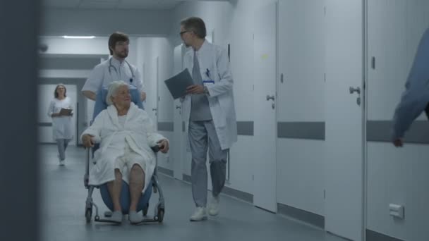 医生推动转乘轮椅 女性病人沿着诊所走廊行走 医生把老年妇女带到手术室或手术室 医护人员和病人在医院走廊 — 图库视频影像