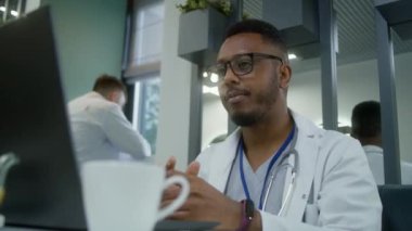 Afro-Amerikalı doktor klinikte oturuyor, dizüstü bilgisayar kullanıyor. Sıhhiyeci hastayla görüntülü konuşma üzerinden görüşüyor, online randevusu var. Hastane veya modern tıp merkezi kantininde tıbbi personel.
