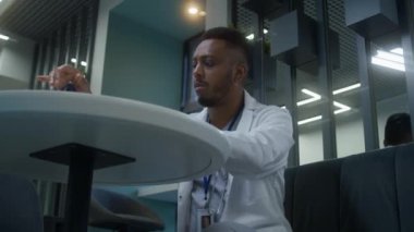 Afrikalı Amerikalı doktor klinikteki kafede oturuyor. Sağlık uzmanı dijital tablet bilgisayarda çalışıyor, hastaların tıbbi test sonuçlarını izliyor. Hastane veya tıp merkezi kafeteryasında tıbbi personel.