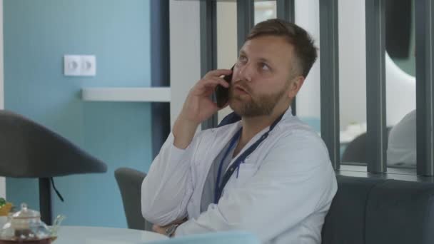男医生坐在诊所咖啡馆休息一下专业医生通过电话与病人交谈 讨论医疗检测结果或治疗 医院或医疗中心食堂的医务人员 — 图库视频影像