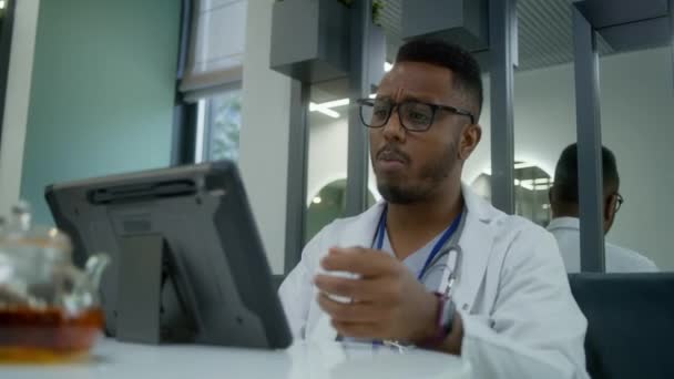 非裔美国医生坐在诊所的咖啡店里 用数字平板电脑 医疗保健专家通过视频聊天咨询病人 有网上预约 医院或医疗中心食堂的医务人员 — 图库视频影像