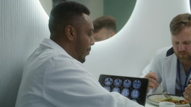 非裔美国医生和同事一起坐在诊所的咖啡店里 医疗保健专家在午餐时交谈 数字平板与Mri脑扫描图像 医院或医疗中心食堂的医务人员 — 图库视频影像