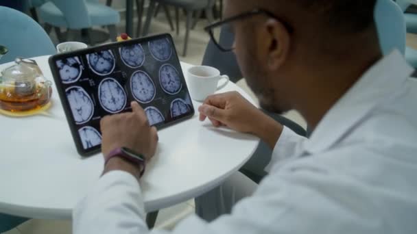 非洲裔美国医生在诊所咖啡店使用数字平板电脑观看Mri或Ct扫描图像 医生喝茶 检查病人的脑部扫描结果 现代医院自助餐厅的医务人员 — 图库视频影像
