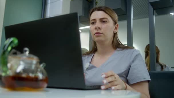 女医生或女护士坐在现代诊所的咖啡店里 使用笔记本电脑 专业医生与病人在线聊天 观看医疗检测结果 上网上网 在医院食堂工作的医务人员 — 图库视频影像