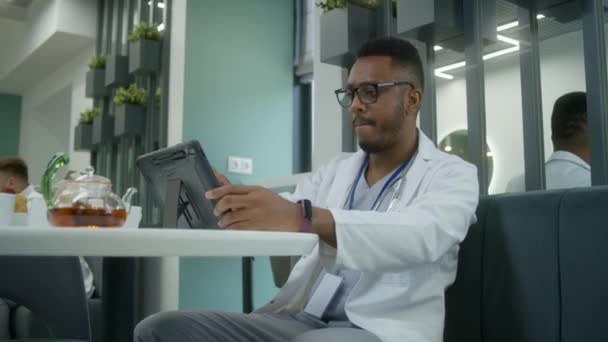 非洲裔美国医生在休息期间在现代诊所咖啡店里使用数字平板电脑 医生喝茶 看病人的体检结果或上网 医院食堂的医务人员 — 图库视频影像