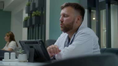 Yetişkin doktor hastane kafesinde oturur ve test sonuçlarını dijital tablet kullanarak görüntülü sohbet yoluyla tartışır. Profesyonel doktor klinik kafeteryasında çalışıyor. Sağlık personeli tıp merkezi kantininde dinleniyor..