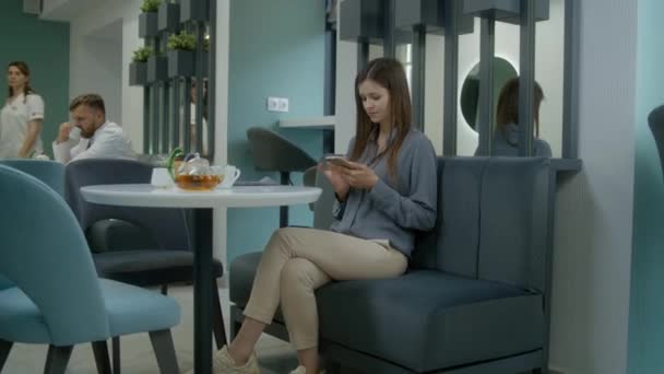 成年妇女坐在现代诊所咖啡厅的沙发上 用电话上网 自助餐女工给女客人送餐 医护人员及病人在医疗中心餐厅共进晚餐 — 图库视频影像