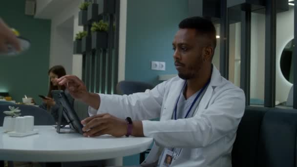 アフリカ系アメリカ人医師がクリニックカフェに座り デジタルタブレットコンピュータで働き 患者の医療検査結果を観察している カフェテリアの労働者は夕食を薬に持って来ます 現代病院の医療スタッフ — ストック動画