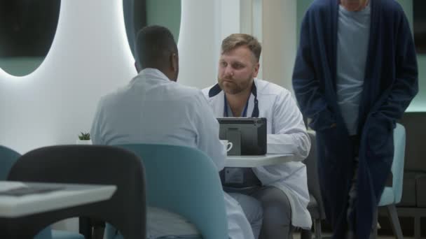 两名多元文化的医生坐在诊所的咖啡店里 讨论工作 男性卫生保健专家与非洲裔美国同事交谈 使用数字平板电脑 医疗人员和医疗中心食堂的病人 — 图库视频影像
