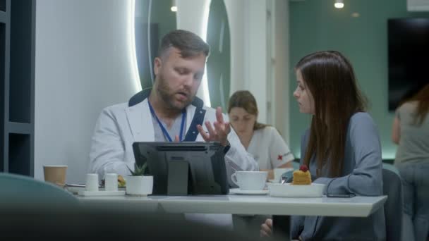 专业医生在医院咖啡店的数字平板电脑上向病人展示了医学检测结果 并对其进行了讨论 女医生在后台吃饭 医院食堂的医务人员 — 图库视频影像