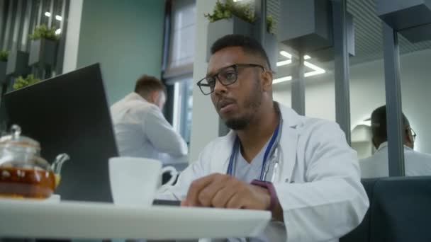 非洲裔美国医生在休息期间在现代诊所的咖啡店里使用笔记本电脑 医生喝茶或咖啡 看病人的体检结果或上网 医院食堂的医务人员 — 图库视频影像