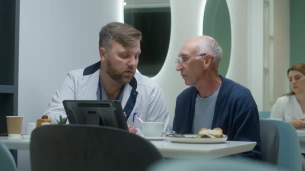 男医生在数码平板电脑上显示老年病人的医疗测试结果 成年医生与老年男子谈论在医疗中心餐厅的治疗 病人在医院咖啡馆吃饭 — 图库视频影像