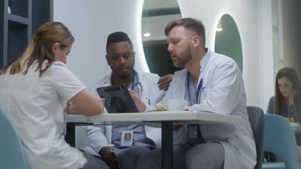 多种族的医生坐在诊所的餐厅里 白人医生吃晚餐 使用数码平板电脑 向他的同事展示测试结果 医护人员在现代医院咖啡馆讨论休息期间的工作 — 图库视频影像
