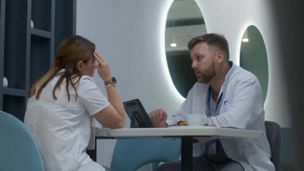 医生和他的同事坐在医院的自助餐厅里 成年医生吃晚餐 在数字平板电脑上检查病人的测试结果 并与护士交谈 医护人员在诊所咖啡店休息 — 图库视频影像