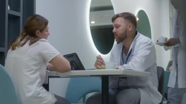 医生在医院餐厅休息 吃饭和说话 非洲医生端来咖啡 请同事们坐下 白种人医生观察数码平板电脑的测试结果 并与其他医生讨论 — 图库视频影像