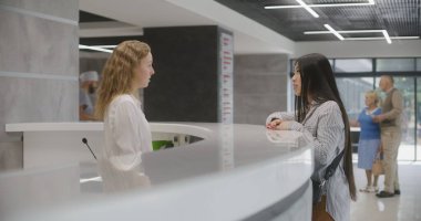 Asyalı kadın klinik lobisindeki resepsiyonun yanında duruyor ve doktorla randevuya geliyor. Bayan yönetici danışmadaki hastayla konuşuyor. Modern hastanede tıbbi personel işi..