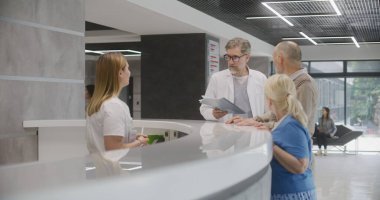 Yaşlı çift tıp merkezinin lobisindeki resepsiyonun yanında durup doktordan randevu alıyorlar. Olgun doktor, danışma masasındaki hastalara danışır. Modern klinikte tıbbi personel.