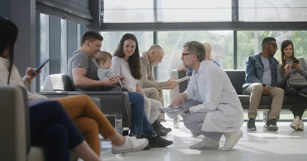 Diverse people sit on couches in clinic lobby area, wait for doctors appointment. Doctor talks to family with little child about medical test results. Waiting room in modern medical center. Healthcare