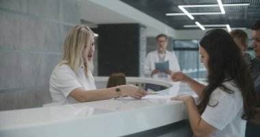 Küçük çocuklu bir aile klinik lobisindeki resepsiyonun yanında duruyor. Kadın evrakları dolduruyor, doktorla randevu alıyor. Modern tıp merkezinde tıbbi personel çalışıyor. Sağlık hizmetleri.