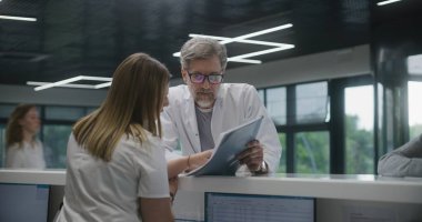 Olgun doktor klinik bekleme odasındaki resepsiyonun yanında duruyor ve hemşireyle işini tartışıyor. Bayan yönetici danışmadaki meslektaşıyla konuşuyor. Modern hastanede tıbbi personel işi..