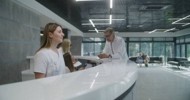 Bayan yönetici klinik lobisindeki resepsiyonda duruyor. Çekici kadın hastaya danışıyor, müşterileri bekliyor, gülümsüyor. Tıp personeli modern tıp merkezinde çalışıyor. Sağlık hizmetleri.