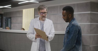 Afro-Amerikalı bir adam randevu sonrası kliniğin lobisinde doktorla birlikte duruyor. Doktor hastayla konuşur, tıbbi muayene ya da test sonuçları gösterir. Tıp merkezinde çalışıyorum. Sağlık hizmetleri sistemi.