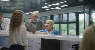 Klinik lobisindeki resepsiyon masasının yanında yaşlı çift duruyor. Doktorla randevuya gelin. Kadın resepsiyonist danışmadaki hastalarla konuşuyor. Modern hastanede tıbbi personel işi..