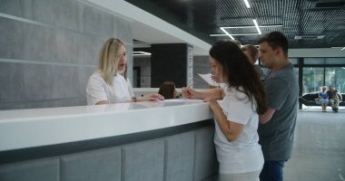 Küçük çocuklu bir aile klinik lobisindeki resepsiyonun yanında duruyor. Kadın evrakları dolduruyor, doktorla randevu alıyor. Modern tıp merkezinde tıbbi personel çalışıyor. Sağlık hizmetleri.