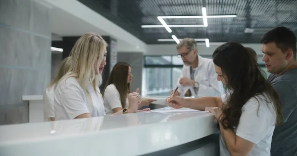 stock image Family with little child stands near reception desk in hospital lobby. Woman fills out documents and papers, makes appointment with doctor. Medical staff work in modern medical center. Health care.