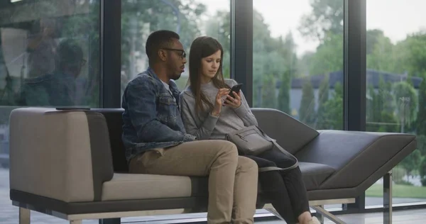 Multiethnic couple sits on sofa in clinic lobby area. Man and woman wait for appointment with doctor or medical test results, surf internet on phone. Waiting area in modern medical center. Healthcare.