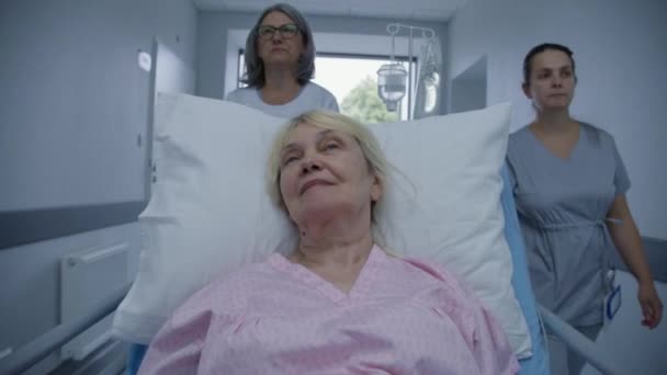 成熟的女医生和年轻的护士把老年病人带到医疗中心 老太婆躺在血淋淋的轮床上 和医生谈论手术的事 在现代医院工作的医务人员 — 图库视频影像