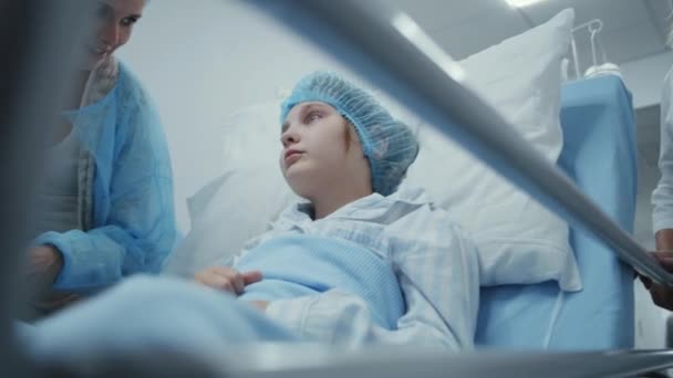 母亲与女儿交谈并牵着手 在动手术前 年轻姑娘戴着医疗帽坐在格尔内 医生把病人送进医疗中心走廊 在现代医院工作的医务人员 — 图库视频影像