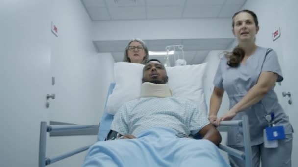 受伤的非裔美国人躺在格尔内的颈部支撑架上 手握静脉导管 还有脉动血压计 成熟的医生和护士将病人运送到医疗中心走廊尽头的病房 — 图库视频影像