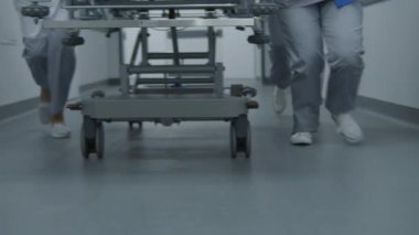 Doktorlar ve sağlık görevlileri koşar, ağır yaralı hastayla birlikte sedyeyi ameliyathaneye iterler. Sıhhiyeciler insan hayatını kurtarır. Tıbbi personel bacaklarını kapatın. Parlak tıbbi tesis koridoru. Yavaş çekim.