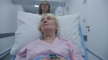Kadın doktor, yaşlı hastayı klinik koridorunda sedyeye itti. Yaşlı kadın sedyede kan damlası ve nabız oksimetresiyle yatıyor. İleride yapılacak ameliyatlar hakkında doktorla konuşuyor. Hastanede çalışan tıbbi personel..