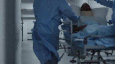 Doktorlar, hemşireler ve sağlık görevlileri hastayla birlikte sedyeyi ameliyathaneye götürün. Boyunluk takmış Afro-Amerikalı bir adam sedyede yatıyor. Acil serviste çok ırklı çalışanlar var. Ağır çekim. Arka plan.